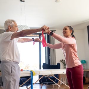 Biracial female physiotherapist with caucasian senior man stretching resistance band at home Assisting unaltered physical therapy exercise strength healthcare retirement training recovery