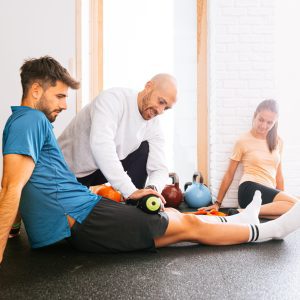 Trainer helping man with foam roller on leg in gym near women