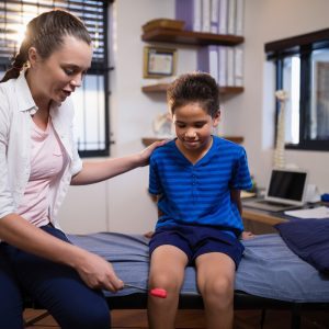 Young female therapist examining boy with reflex hammer on knee while sitting on bed
